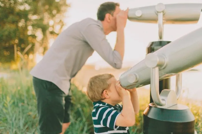 Un adulte et un enfant regardent dans des jumelles à pièces en plein air par une journée ensoleillée. L'adulte se tient derrière l'enfant, tous deux orientés dans la même direction, entourés de verdure.