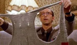 Un homme portant des lunettes et un costume brandit un vêtement tricoté gris avec de grandes emmanchures, le regardant avec une expression de surprise ou de confusion. Une lampe et un papier peint à motifs se trouvent à l'arrière-plan.