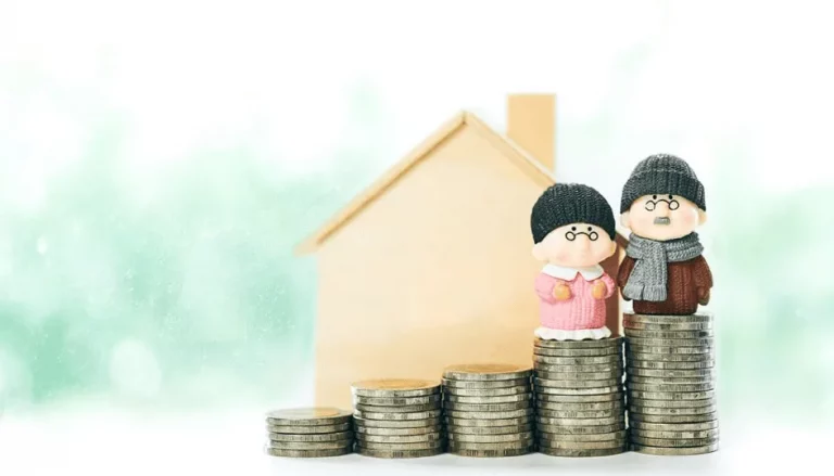 Deux figurines âgées portant des vêtements d'hiver se tiennent au-dessus de pièces de monnaie empilées, avec une maquette de maison en bois à l'arrière-plan, symbolisant l'épargne-retraite et la planification financière pour la vieillesse.