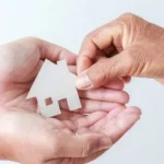 Deux mains tiennent une petite maison en bois découpée, tandis qu'une troisième la place délicatement dans les paumes ouvertes, symbolisant le don ou le transfert d'une maison.