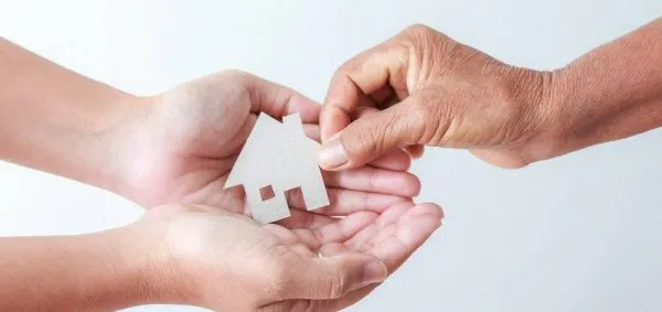 Deux mains tiennent une petite maison en bois découpée, tandis qu'une troisième la place délicatement dans les paumes ouvertes, symbolisant le don ou le transfert d'une maison.