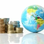 Des piles de pièces de monnaie assorties sont disposées à côté d'un petit globe sur une surface blanche réfléchissante, symbolisant la finance et l'économie mondiales.