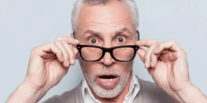 Un homme âgé aux cheveux et à la barbe grisonnants, portant des lunettes et un pull léger, soulève ses lunettes et prend un air surpris, la bouche ouverte et les yeux écarquillés, sur un fond clair.