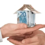 Une maquette de maison en billets d'euros est tenue entre deux mains, symbolisant une transaction ou un échange financier, sur un fond blanc uni.