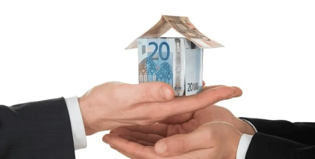 Une maquette de maison en billets d'euros est tenue entre deux mains, symbolisant une transaction ou un échange financier, sur un fond blanc uni.