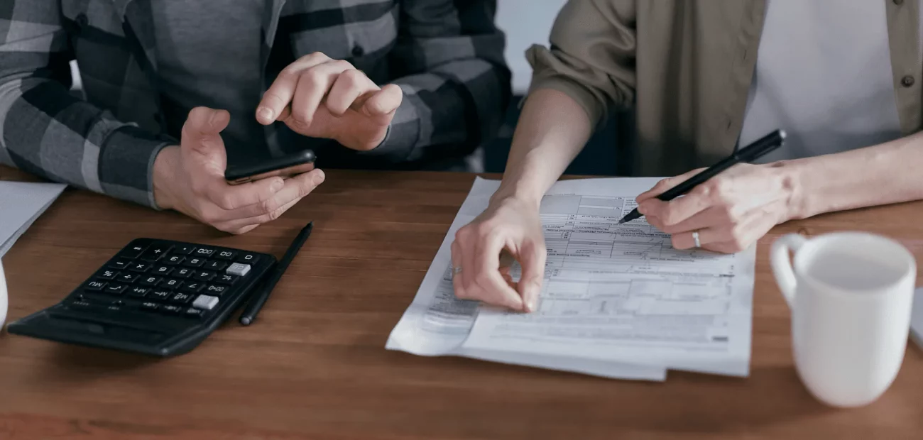 Deux personnes sont assises à une table et travaillent sur des documents. L'une tient un téléphone, l'autre pointe un document avec un stylo. Une calculatrice et un mug blanc se trouvent également sur la table en bois.