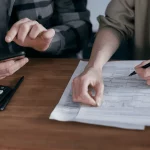 Deux personnes sont assises à une table et travaillent sur des documents. L'une tient un téléphone, l'autre pointe un document avec un stylo. Une calculatrice et un mug blanc se trouvent également sur la table en bois.