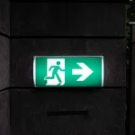 Un panneau de sortie de secours vert avec une silhouette en mouvement et une flèche pointant vers la droite est fixé sur un mur sombre, indiquant la direction de la sortie la plus proche.