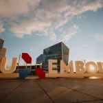 De grandes lettres sculpturales épelant LUXEMBOURG se dressent sur une place au coucher du soleil, avec des bâtiments modernes en verre et un ciel partiellement nuageux en arrière-plan.