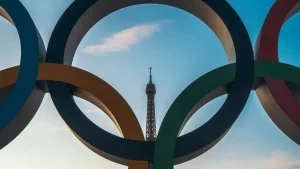 La Tour Eiffel est vue à travers les anneaux du logo olympique, avec un ciel bleu en arrière-plan. Les anneaux olympiques sont grands et colorés, encadrant le symbole de Paris au centre.