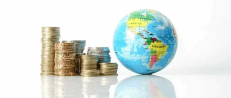 Un petit globe terrestre repose sur une surface réfléchissante à côté de plusieurs piles de pièces de monnaie de différentes hauteurs, symbolisant la finance mondiale ou l'économie mondiale.