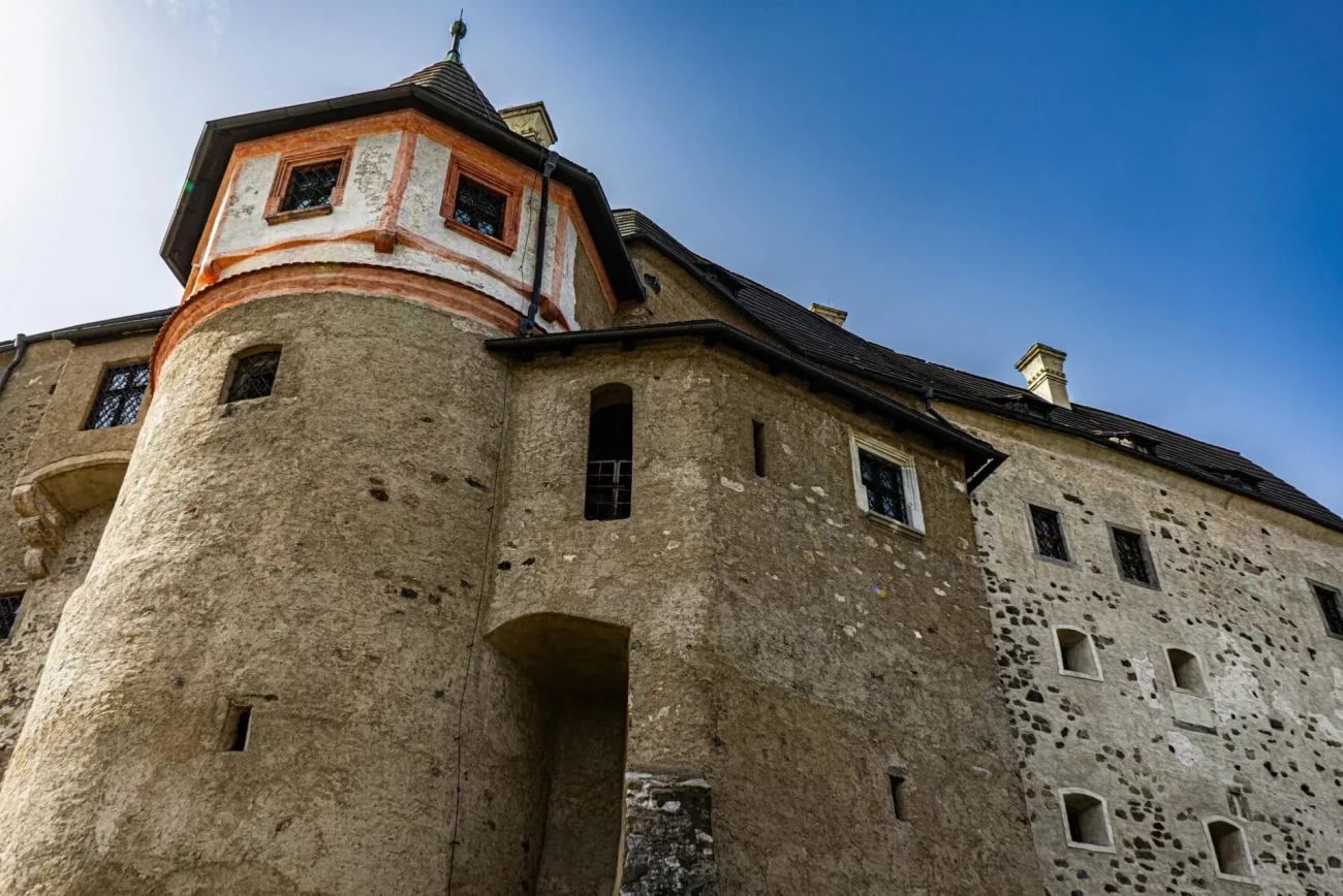 Vue en contre-plongée d'un château historique en pierre aux murs rugueux et texturés, aux petites fenêtres et à la tourelle ronde aux accents peints en rouge et blanc, sur fond de ciel bleu clair.
