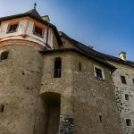Vue en contre-plongée d'un château historique en pierre aux murs rugueux et texturés, aux petites fenêtres et à la tourelle ronde aux accents peints en rouge et blanc, sur fond de ciel bleu clair.