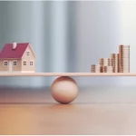 Une petite maquette de maison et des piles de pièces de monnaie sont placées en équilibre sur les extrémités opposées d'une planche de bois, qui repose sur une boule ronde en bois, symbolisant l'équilibre entre la propriété et l'argent.