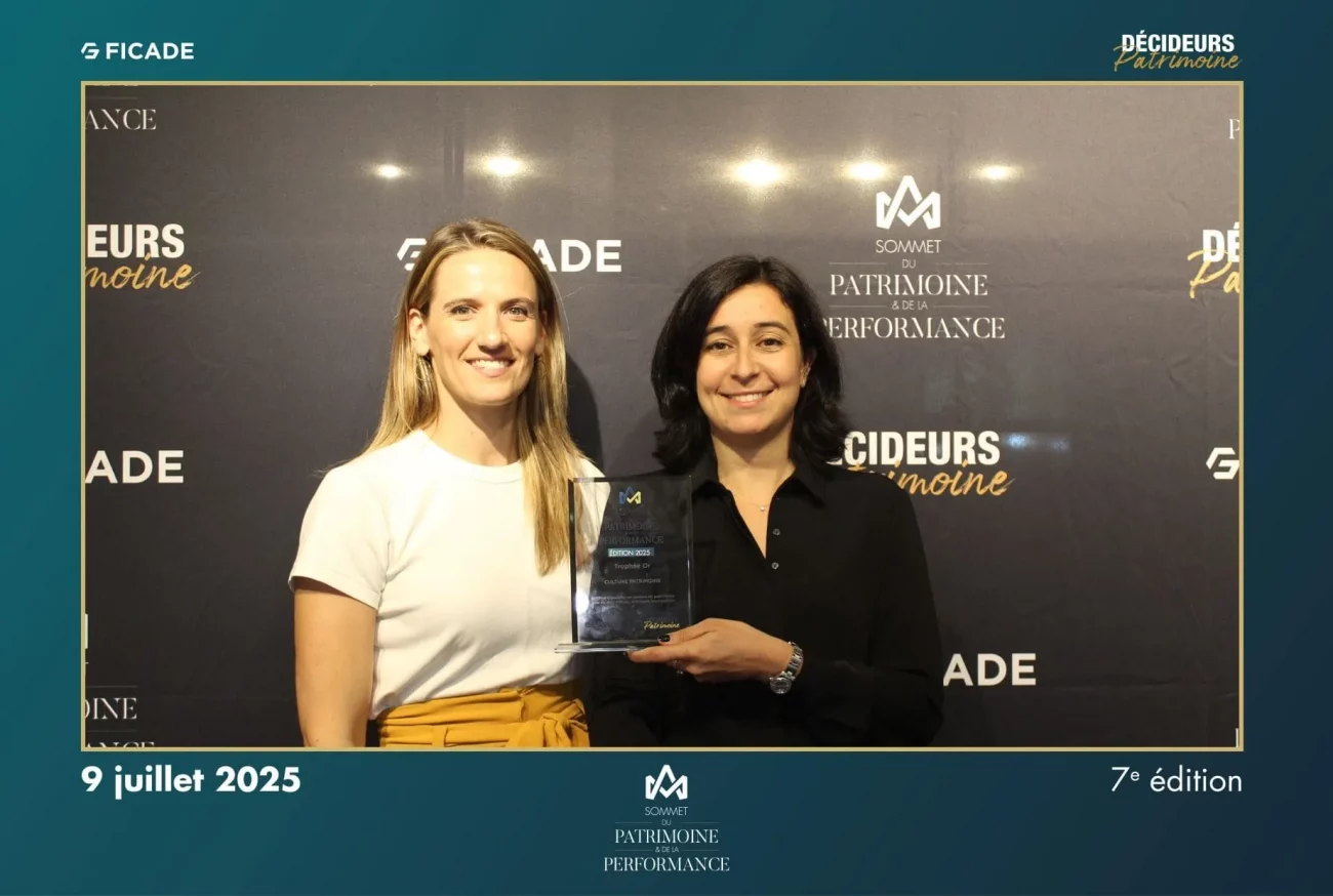 Deux femmes sourient devant une toile de fond. L'une d'elles, à droite, tient un prix en verre. La toile de fond affiche des logos et du texte pour un événement nommé Décideurs Patrimoine le 9 juillet 2025.