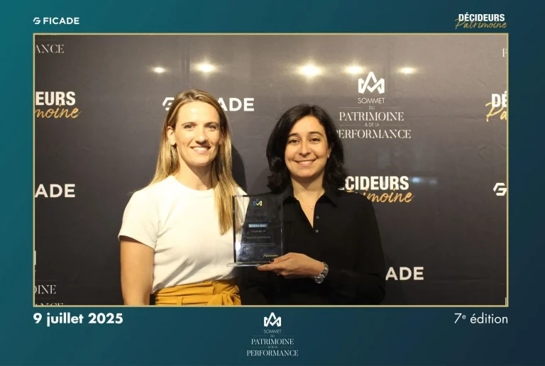 Deux femmes sourient devant une toile de fond. L'une d'elles, à droite, tient un prix en verre. La toile de fond affiche des logos et du texte pour un événement nommé Décideurs Patrimoine le 9 juillet 2025.