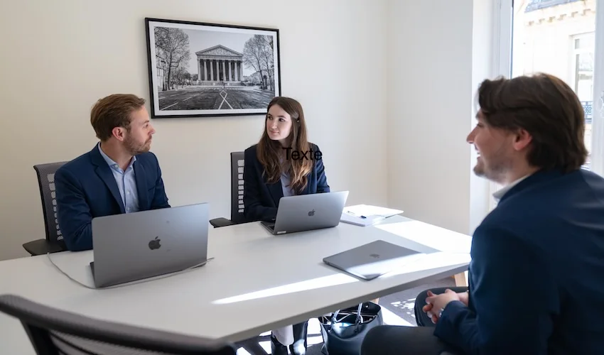 Dans un bureau lumineux, trois personnes sont assises autour d'une table blanche, chacune munie d'un ordinateur portable. Elles semblent être en train de discuter. Une photo encadrée en noir et blanc d'un bâtiment classique est accrochée au mur derrière eux.