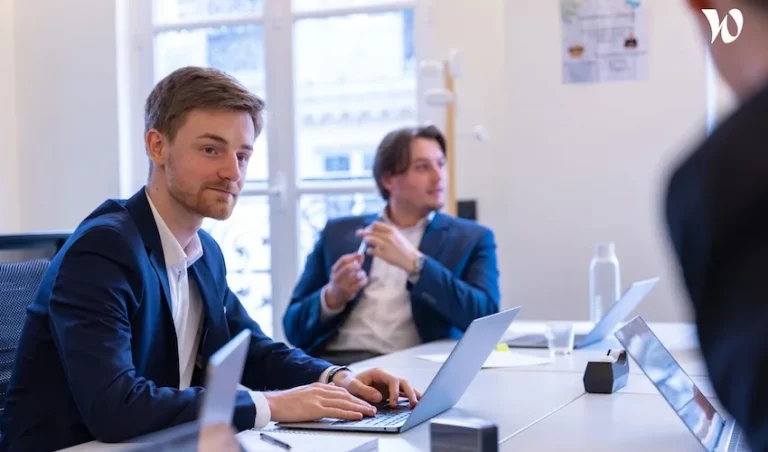 Deux hommes en costume d'affaires sont assis à une table de conférence avec des ordinateurs portables dans un bureau lumineux. L'un d'eux fait face à la caméra, écoutant attentivement, tandis que l'autre regarde ailleurs, faisant des gestes pendant la discussion.