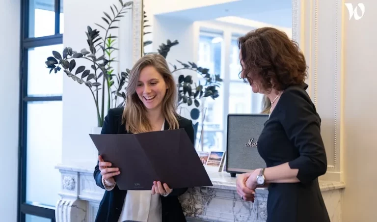 Deux femmes se tiennent dans un bureau, souriant et regardant ensemble des documents. Elles sont près d'une cheminée blanche avec une plante, des photos et un haut-parleur sur le manteau. L'atmosphère semble professionnelle et amicale.