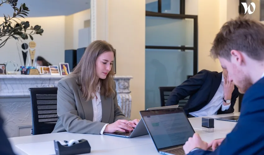 Trois personnes sont assises à une table de conférence et travaillent sur des ordinateurs portables. Une femme vêtue d'un blazer tape à la machine, tandis que deux hommes, dont l'un a le visage dans la main, se concentrent sur leur écran. Le cadre est celui d'un bureau moderne et lumineux.